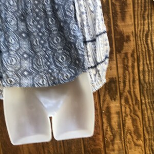 Puede incluir: Una blusa blanca y azul estampada con mangas largas. La blusa presenta un escote cuadrado y un dise&ntilde;o detallado en el corpi&ntilde;o. Las mangas tienen un estampado de rayas con detalles en azul oscuro. La prenda se exhibe en un maniqu&iacute; blanco.