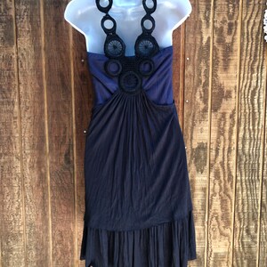 Op de afbeelding: Een marineblauwe jurk met een halterhalslijn, weergegeven op een witte mannequin. De halter heeft zwarte cirkelvormige en bloemenhaakdetails. De jurk heeft een geplooid lijfje en een gefranjerde zoom, waardoor een vloeiend silhouet ontstaat.