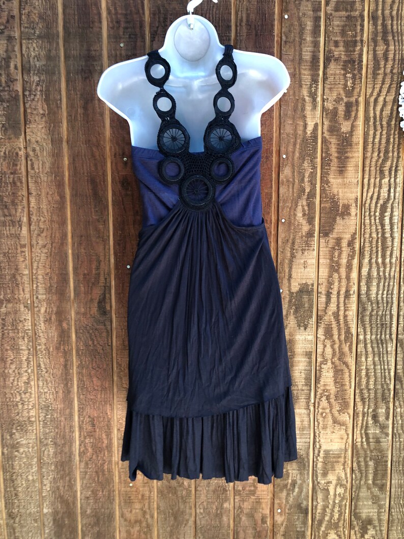 Op de afbeelding: Een marineblauwe jurk met een halterhalslijn, weergegeven op een witte mannequin. De halter heeft zwarte cirkelvormige en bloemenhaakdetails. De jurk heeft een geplooid lijfje en een gefranjerde zoom, waardoor een vloeiend silhouet ontstaat.