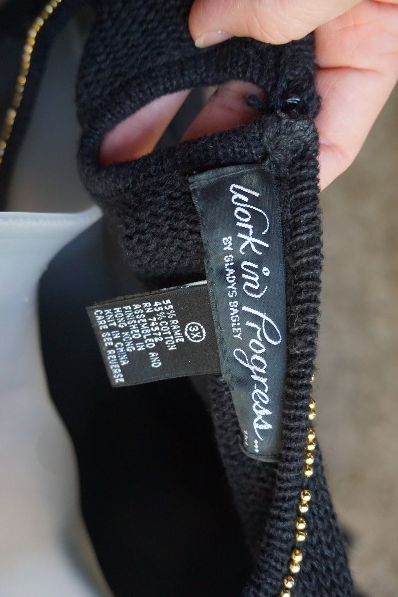 Pode incluir: Camisola preta com detalhes de contas douradas e uma etiqueta preta que diz "Work in Progress... by Sladys Bagley". A etiqueta tamb&eacute;m indica que a pe&ccedil;a &eacute; feita de 55% ramie e 45% algod&atilde;o. A etiqueta tamb&eacute;m indica que a pe&ccedil;a foi montada e acabada em Hong Kong, tricotada na China e tem um tamanho 3X.