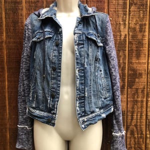 May include: A blue denim jacket with a distressed look, featuring a hood and knit sleeves. The jacket has button closures and is displayed on a mannequin against a wooden background.