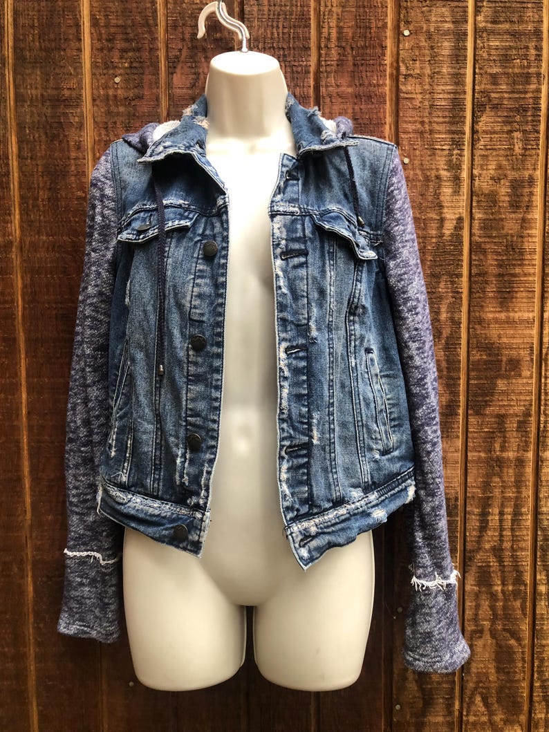 May include: A blue denim jacket with a distressed look, featuring a hood and knit sleeves. The jacket has button closures and is displayed on a mannequin against a wooden background.