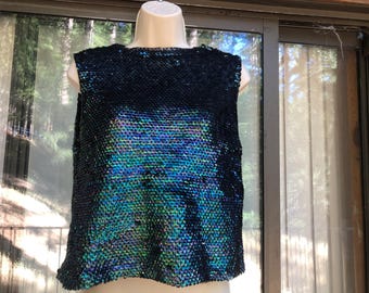 May include: A sleeveless top covered in iridescent sequins. The sequins shift in color, displaying shades of blue, green, and purple. The top is displayed on a mannequin and appears to be a party or evening wear garment.