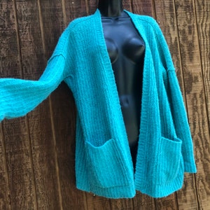 May include: A teal blue open-front cardigan with long sleeves and two pockets. The cardigan is made of a soft, fuzzy material.