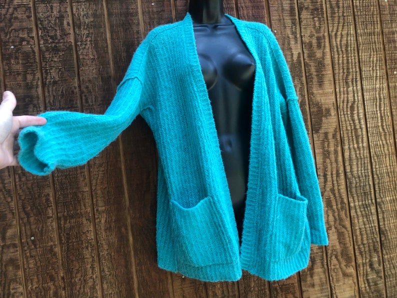 May include: A teal blue open-front cardigan with long sleeves and two pockets. The cardigan is made of a soft, fuzzy material.
