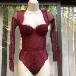 May include: Burgundy lace bodysuit with long sheer sleeves. The lingerie features a sweetheart neckline and a delicate floral lace pattern. The garment is displayed on a white mannequin against a window with a natural background.