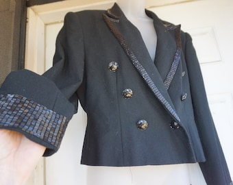 May include: Black blazer with decorative trim on the lapels, cuffs, and collar. The blazer has a double-breasted design with large, round buttons. The trim appears to be a textured, dark material.