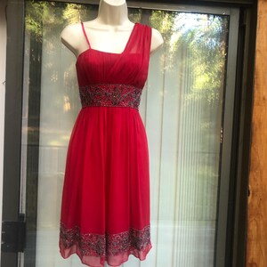 K&ouml;nnte beinhalten: Ein rotes Cocktailkleid mit One-Shoulder-Design und einem bestickten Taillenband. Das Kleid hat einen knielangen Saum und einen zarten, flie&szlig;enden Stoff. Das Kleid wird auf einer Schaufensterpuppe pr&auml;sentiert.