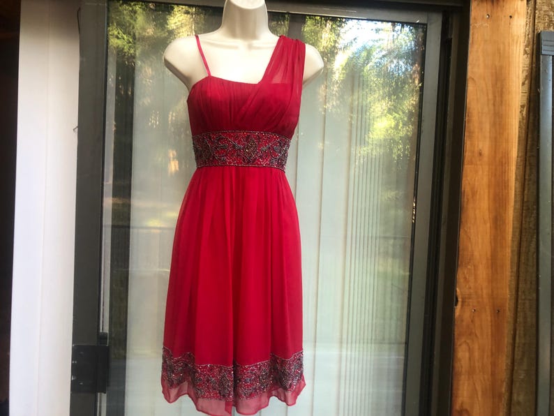 K&ouml;nnte beinhalten: Ein rotes Cocktailkleid mit One-Shoulder-Design und einem bestickten Taillenband. Das Kleid hat einen knielangen Saum und einen zarten, flie&szlig;enden Stoff. Das Kleid wird auf einer Schaufensterpuppe pr&auml;sentiert.