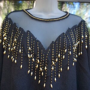 Pode incluir: Camisola preta com decote de malha transparente e detalhe de franjas de contas douradas.