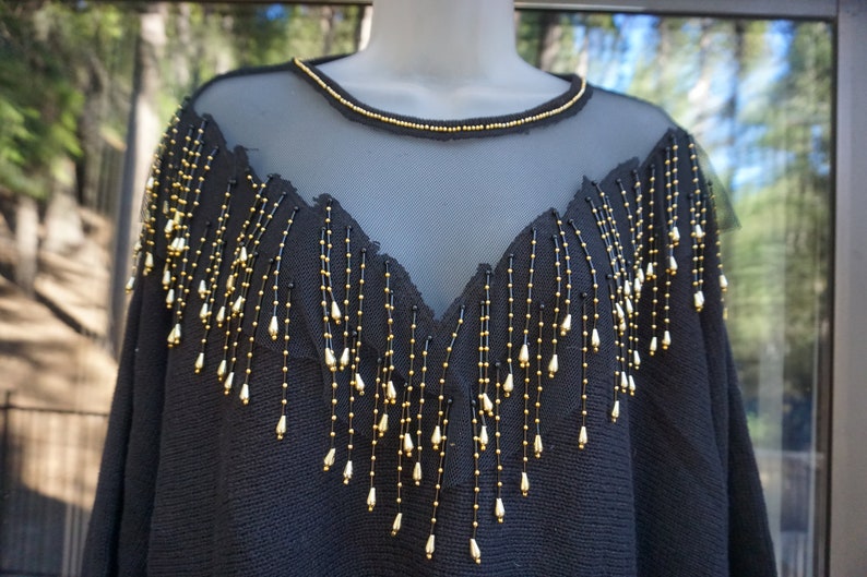 Pode incluir: Camisola preta com decote de malha transparente e detalhe de franjas de contas douradas.