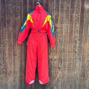 May include: A vintage red ski suit with yellow and teal detailing. The one-piece suit has a belted waist, a high collar, and a zip closure. The suit is hanging on a metal hanger against a wooden backdrop.