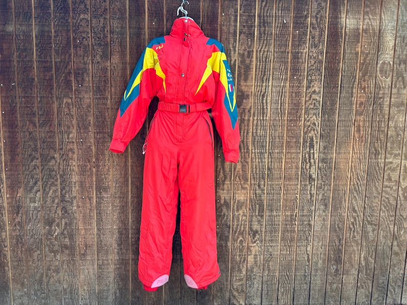 May include: A vintage red ski suit with yellow and teal detailing. The one-piece suit has a belted waist, a high collar, and a zip closure. The suit is hanging on a metal hanger against a wooden backdrop.