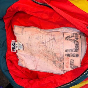 May include: A vintage Fila jacket in red with yellow and green detailing. The interior is red with a large printed label. The jacket features a zipper and a Fila label. The jacket is a retro style.