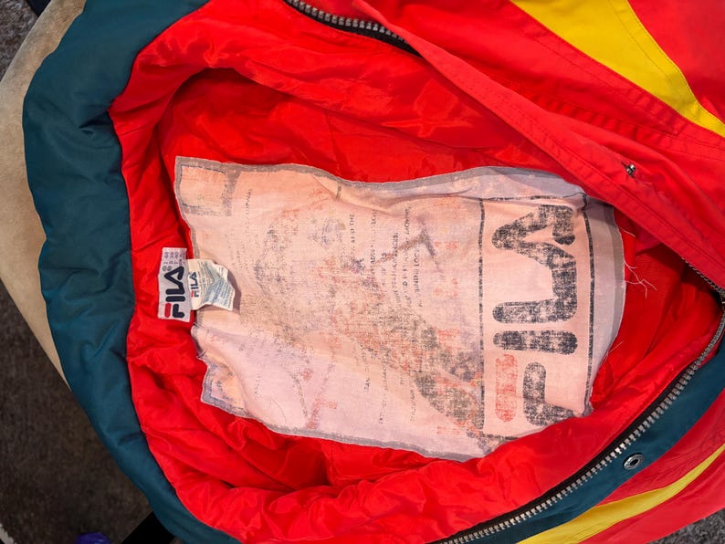 May include: A vintage Fila jacket in red with yellow and green detailing. The interior is red with a large printed label. The jacket features a zipper and a Fila label. The jacket is a retro style.
