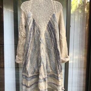 May include: A beige knitted cardigan with a blue and white striped pattern. The cardigan has a long, flowing design and is made of a soft, lightweight material.