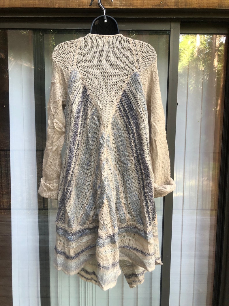 May include: A beige knitted cardigan with a blue and white striped pattern. The cardigan has a long, flowing design and is made of a soft, lightweight material.