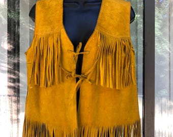 May include: Mustard yellow suede vest with long fringe detailing. The vest has a V-neck and a tie closure at the front. The fringe is along the bottom and the chest area. The vest is displayed on a black mannequin.