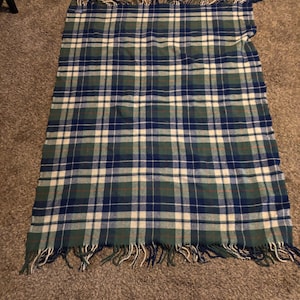 May include: A rectangular throw blanket featuring a plaid pattern in shades of dark blue, forest green, and cream. The fabric has a woven texture and is finished with short, loose fringe along the top and bottom edges of the textile piece.