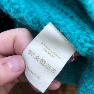 May include: A white tag with a gold label that reads "87% Alpaca/Alpaca 13% Nylon" and care instructions. The tag is attached to a teal fuzzy sweater.