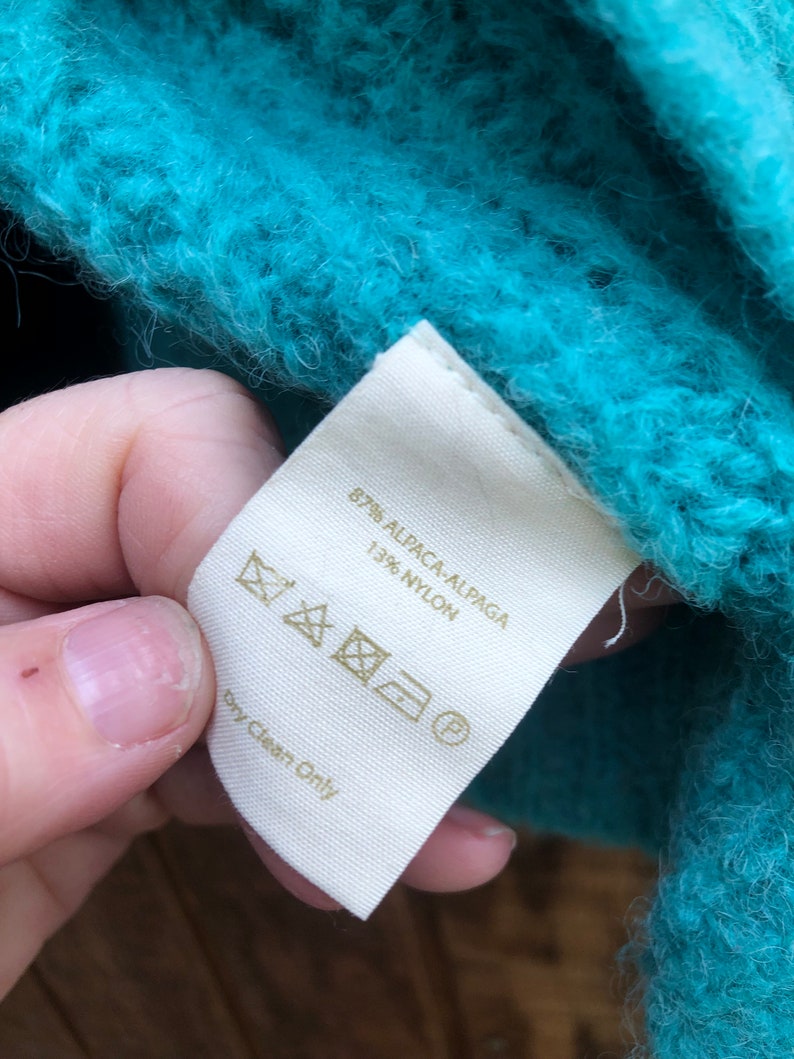 May include: A white tag with a gold label that reads "87% Alpaca/Alpaca 13% Nylon" and care instructions. The tag is attached to a teal fuzzy sweater.