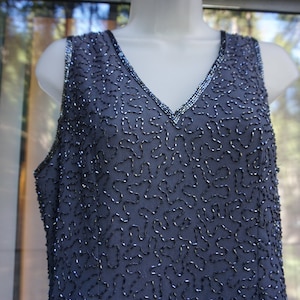 May include: A sleeveless, dark blue dress with a V-neckline. The dress is embellished with silver-colored beads in a flowing, abstract pattern. The neckline and straps are also trimmed with silver beads, adding a touch of sparkle.