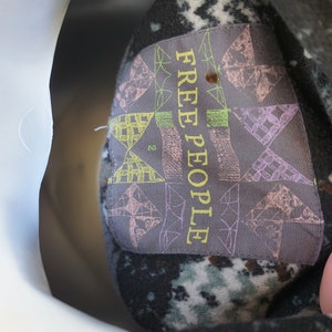 May include: Close-up of a garment tag with the text "FREE PEOPLE" in yellow. The tag is gray with geometric patterns in pink, green, and yellow. The garment has a dark, speckled pattern.