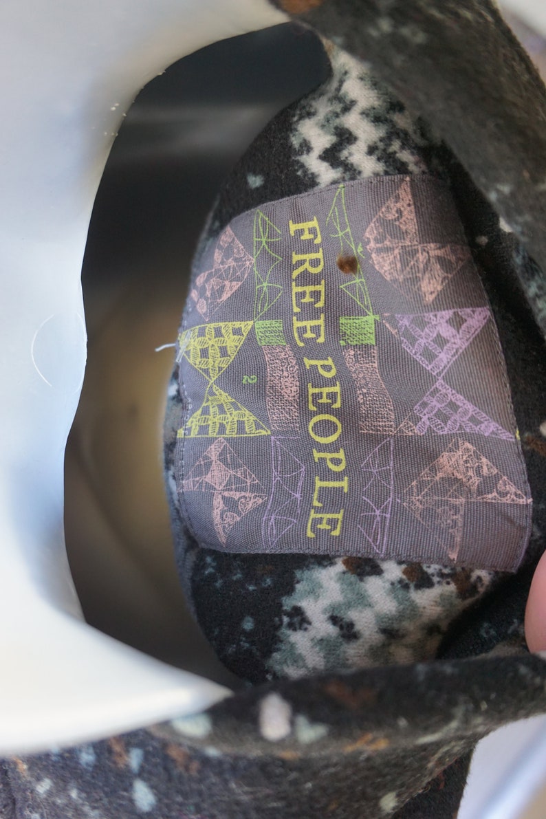 May include: Close-up of a garment tag with the text "FREE PEOPLE" in yellow. The tag is gray with geometric patterns in pink, green, and yellow. The garment has a dark, speckled pattern.