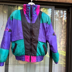 May include: A vintage color-blocked jacket in shades of purple, teal, black, and pink. The jacket features a high collar, a full-length zipper, and elastic cuffs and hem. Geometric shapes are sewn on the sleeves. This retro jacket is a vibrant statement piece.
