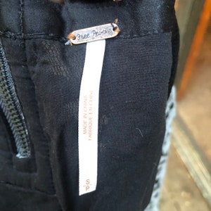 May include: A white tag with black text that reads "Free People" and "Made in China / Fabrique en Chine". The tag is attached to a black garment with a zipper.