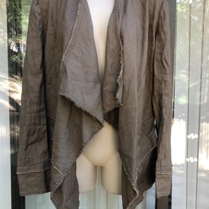 K&ouml;nnte beinhalten: Eine offene Jacke in Taupe mit einem drapierten Design. Die Jacke hat lange &Auml;rmel und ausgefranste Kanten am Kragen und an den Frontpartien. Der Stoff scheint ein leichter Leinenstoff oder ein &auml;hnliches Material zu sein.