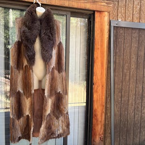 May include: A long, sleeveless vest featuring a patchwork design of brown and tan fur panels. The garment includes a large, dark brown fur collar that extends down the front, creating a textured, layered appearance with a vertical, open silhouette.