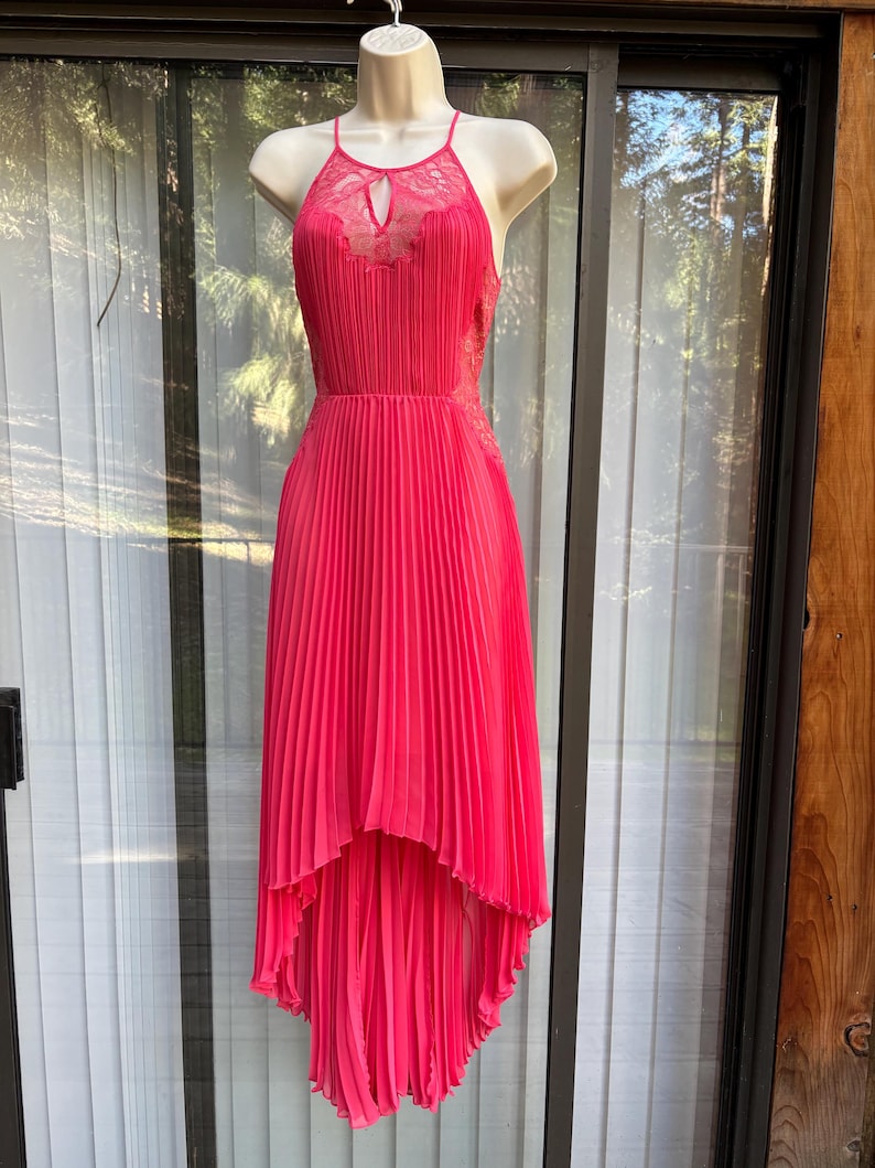 Puede incluir: Un vestido rosa brillante con cuello halter, falda plisada y dobladillo asim&eacute;trico. El corpi&ntilde;o presenta un detalle de ojo de cerradura y detalles de encaje. El vestido se exhibe en un maniqu&iacute;.