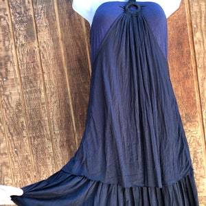 Op de afbeelding: Een marineblauwe halterjurk op een witte mannequin. De jurk heeft een donkerblauwe lijfje met een cirkelvormig accent en een vloeiende, geplooide rok. De zoom heeft een gefranjerde rand. De jurk wordt getoond tegen een houten achtergrond.