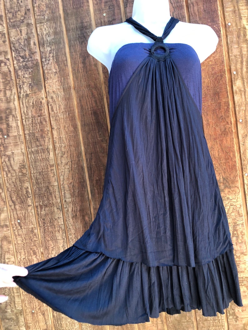 Op de afbeelding: Een marineblauwe halterjurk op een witte mannequin. De jurk heeft een donkerblauwe lijfje met een cirkelvormig accent en een vloeiende, geplooide rok. De zoom heeft een gefranjerde rand. De jurk wordt getoond tegen een houten achtergrond.