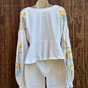 May include: White blouse with long, billowing sleeves and intricate floral embroidery in yellow, blue, and green. The blouse features a scoop neckline and a peplum waist, with tie details at the cuffs. The garment is displayed on a white mannequin against a wooden background.