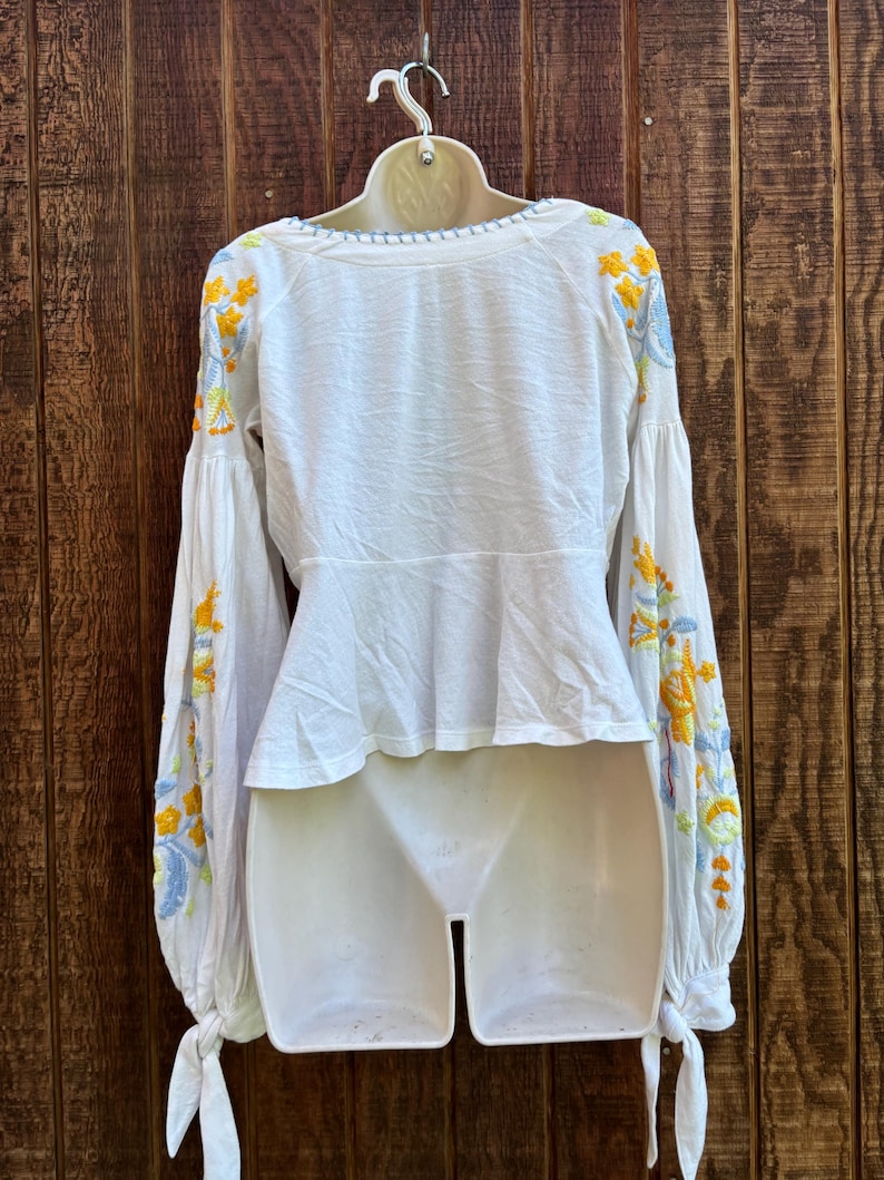 May include: White blouse with long, billowing sleeves and intricate floral embroidery in yellow, blue, and green. The blouse features a scoop neckline and a peplum waist, with tie details at the cuffs. The garment is displayed on a white mannequin against a wooden background.