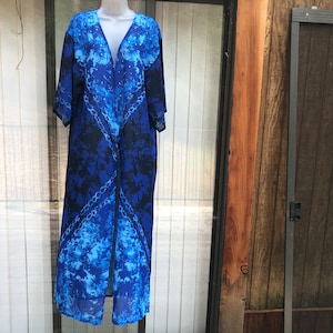 May include: A long, flowing blue kimono-style robe with a V-neck. The robe features a tie-dye pattern in shades of blue and black. The garment is displayed on a mannequin, showcasing its open front and long sleeves.