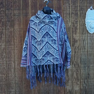 May include: A blue and white patterned poncho with a cowl neck and fringe. The poncho has a geometric design with red accents and is displayed on a black mannequin head against a wooden background.