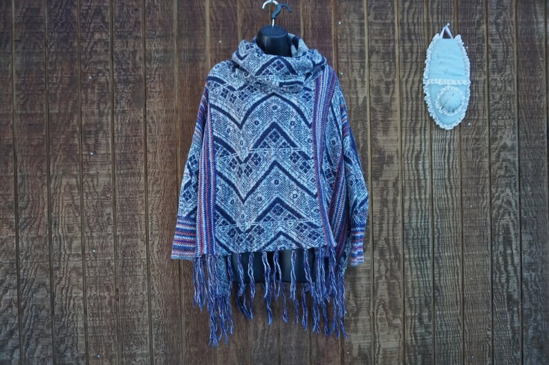 May include: A blue and white patterned poncho with a cowl neck and fringe. The poncho has a geometric design with red accents and is displayed on a black mannequin head against a wooden background.
