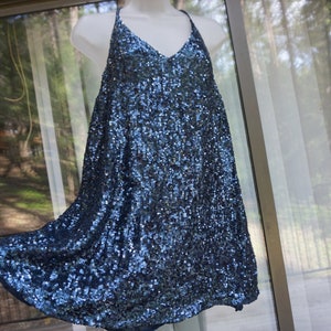 K&ouml;nnte beinhalten: Ein marineblaues Paillettenkleid mit V-Ausschnitt und Spaghettitr&auml;gern. Das Kleid wird auf einer wei&szlig;en Schaufensterpuppe pr&auml;sentiert und hat einen flie&szlig;enden, ausgestellten Rock. Die Pailletten erzeugen einen schimmernden Effekt.