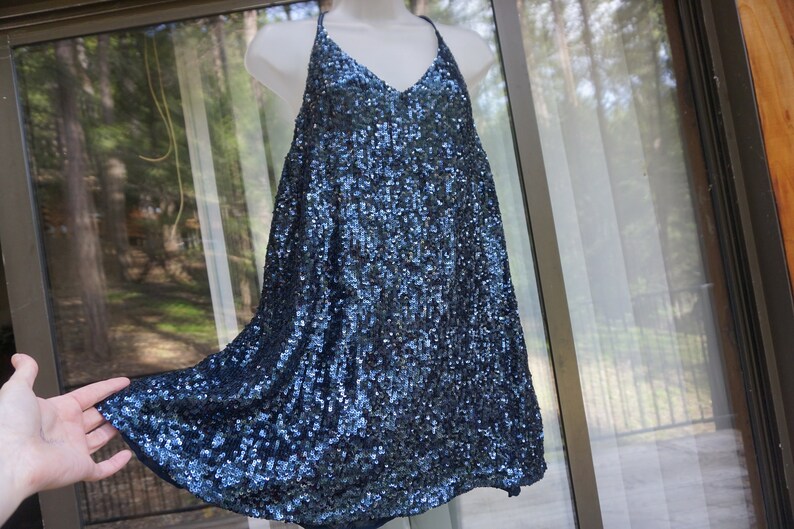 K&ouml;nnte beinhalten: Ein marineblaues Paillettenkleid mit V-Ausschnitt und Spaghettitr&auml;gern. Das Kleid wird auf einer wei&szlig;en Schaufensterpuppe pr&auml;sentiert und hat einen flie&szlig;enden, ausgestellten Rock. Die Pailletten erzeugen einen schimmernden Effekt.