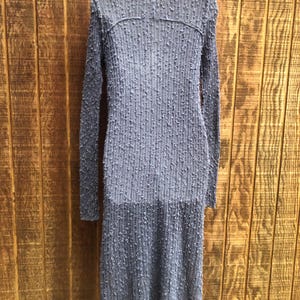 K&ouml;nnte beinhalten: Ein langes, strukturiertes, graues Kleid mit langen &Auml;rmeln h&auml;ngt an einem Kleiderb&uuml;gel. Das Kleid hat einen hohen Ausschnitt und ein strukturiertes, gepunktetes Muster im gesamten Stoff. Das Kleid besteht aus einem leichten Material und hat einen leichten Saum.