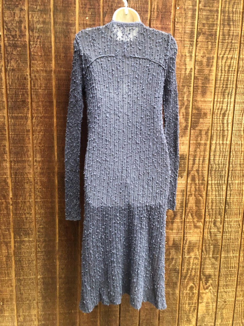 K&ouml;nnte beinhalten: Ein langes, strukturiertes, graues Kleid mit langen &Auml;rmeln h&auml;ngt an einem Kleiderb&uuml;gel. Das Kleid hat einen hohen Ausschnitt und ein strukturiertes, gepunktetes Muster im gesamten Stoff. Das Kleid besteht aus einem leichten Material und hat einen leichten Saum.
