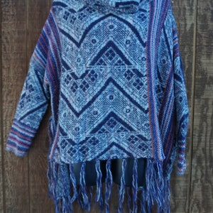 May include: A blue and white patterned poncho with a cowl neck and fringe. The poncho features a geometric design in shades of blue and white, with red accents. The fringe is a mix of blue and white threads. The garment is displayed on a black mannequin.