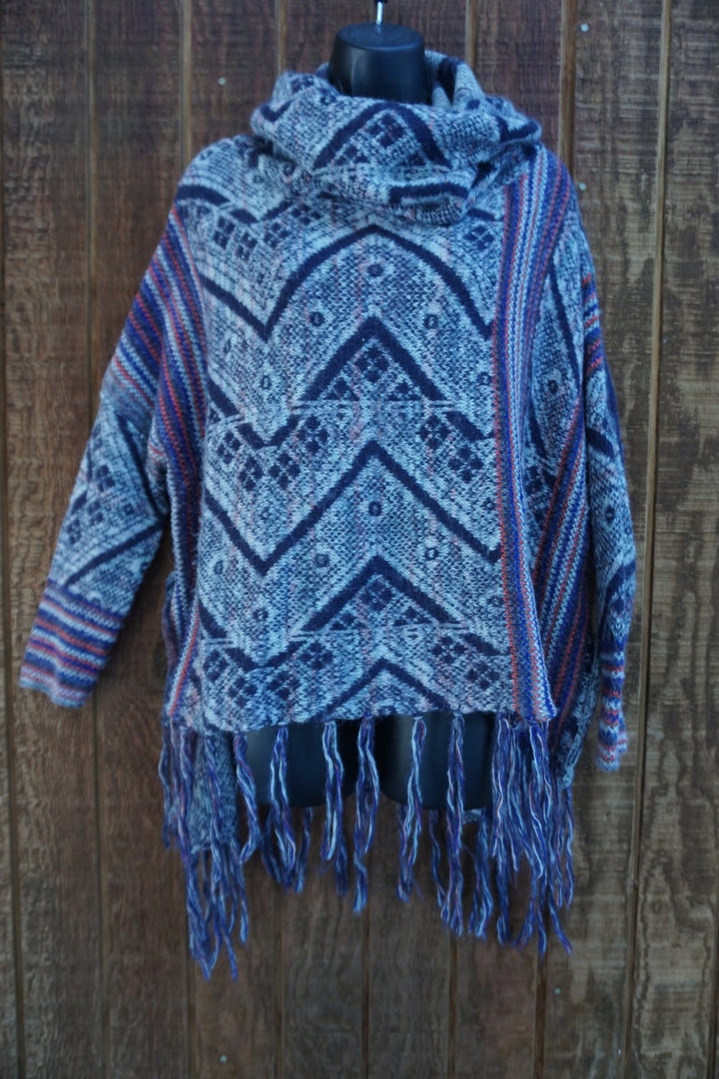 May include: A blue and white patterned poncho with a cowl neck and fringe. The poncho features a geometric design in shades of blue and white, with red accents. The fringe is a mix of blue and white threads. The garment is displayed on a black mannequin.