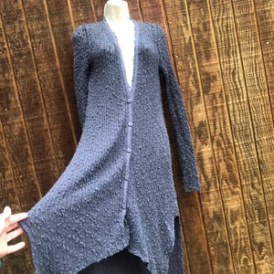 K&ouml;nnte beinhalten: Langer, gekn&ouml;pfter Cardigan aus strukturiertem, dunkelblauem Stoff. Der Cardigan hat lange &Auml;rmel und einen High-Low-Saum. Das Kleidungsst&uuml;ck wird auf einer Schaufensterpuppe vor einem Holzgrund gezeigt.