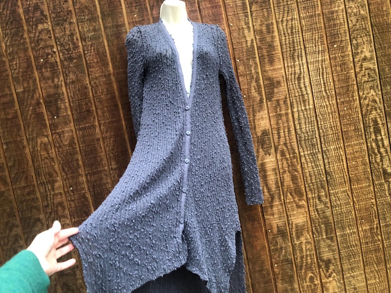 K&ouml;nnte beinhalten: Langer, gekn&ouml;pfter Cardigan aus strukturiertem, dunkelblauem Stoff. Der Cardigan hat lange &Auml;rmel und einen High-Low-Saum. Das Kleidungsst&uuml;ck wird auf einer Schaufensterpuppe vor einem Holzgrund gezeigt.
