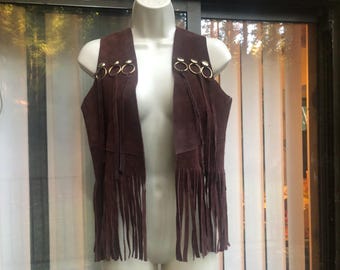 May include: A burgundy suede vest with a fringed hem and gold-tone ring accents. The open-front vest is displayed on a mannequin. The vest has a bohemian style.