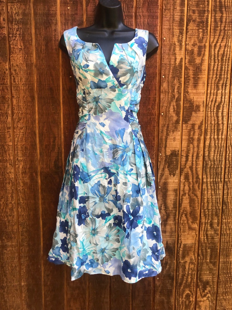May include: Sleeveless floral dress in shades of blue, white, and gray. The dress features a V-neckline and a fitted bodice with a pleated skirt. The floral pattern includes large and small blooms.